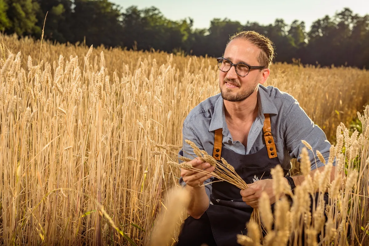 Vor zehn Jahren hat Martin Mayer die Bäckerei Vuaillat übernommen und in eine Aktiengesellschaft umgewandelt. Martin Mayer kniet in einem Feld mit goldfarbenem reifem Weizen. Er trägt eine Schürze.